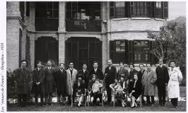 Photo en noir et blanc d'un groupe de 16 personnes (hommes, femmes et enfants) pose devant un bâtiment en brique à fenêtres voûtées et volets noirs, probablement une résidence ou une institution. Les adultes portent des manteaux et costumes formels des années 1920, tandis que les enfants sont assis sur des chaises au premier rang. Des arbres encadrent la scène, suggérant un jardin ou une cour.